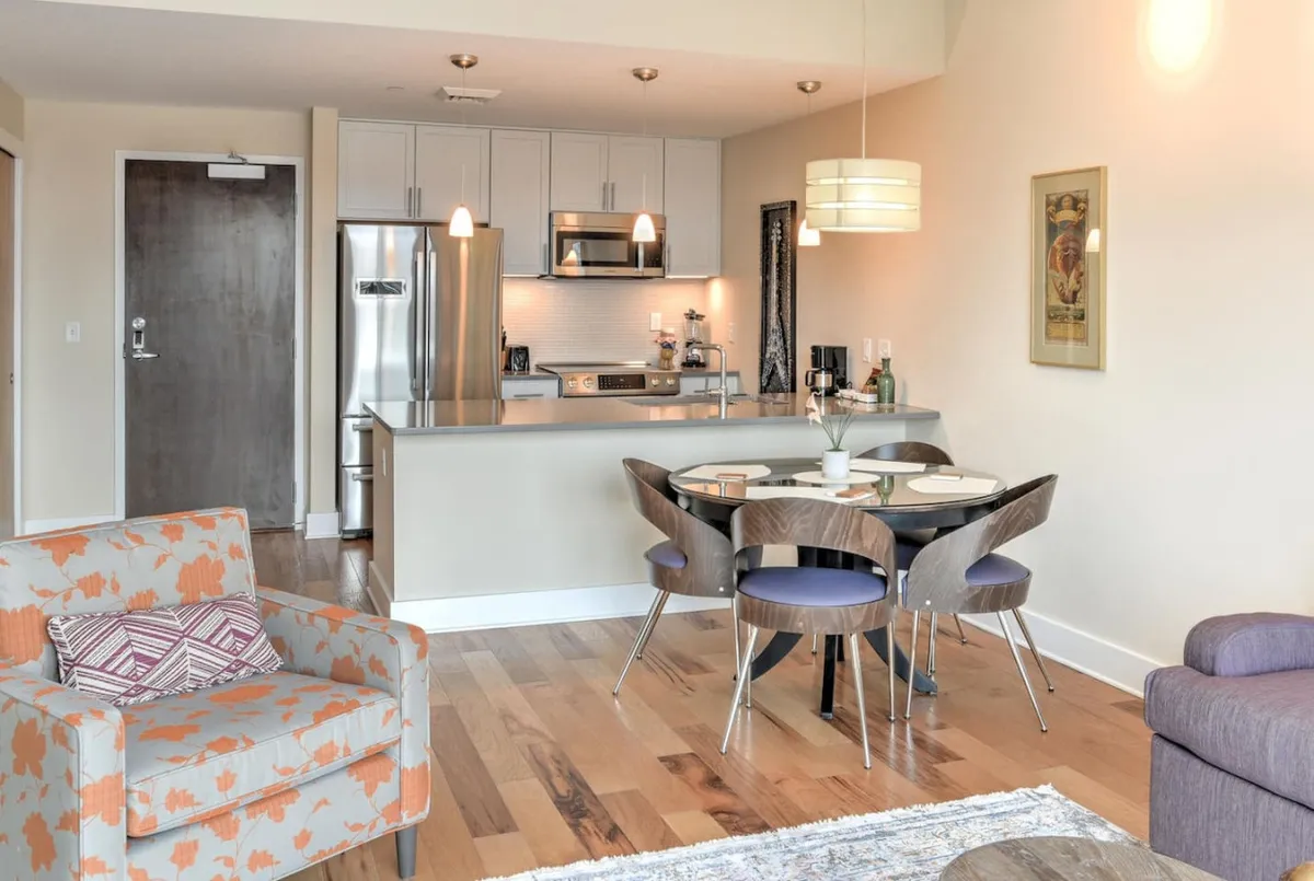 A modern open-plan space with a dining table, kitchen area, sofa, and a decorative chair. The environment is contemporary and functional, ideal for both relaxation and entertaining.