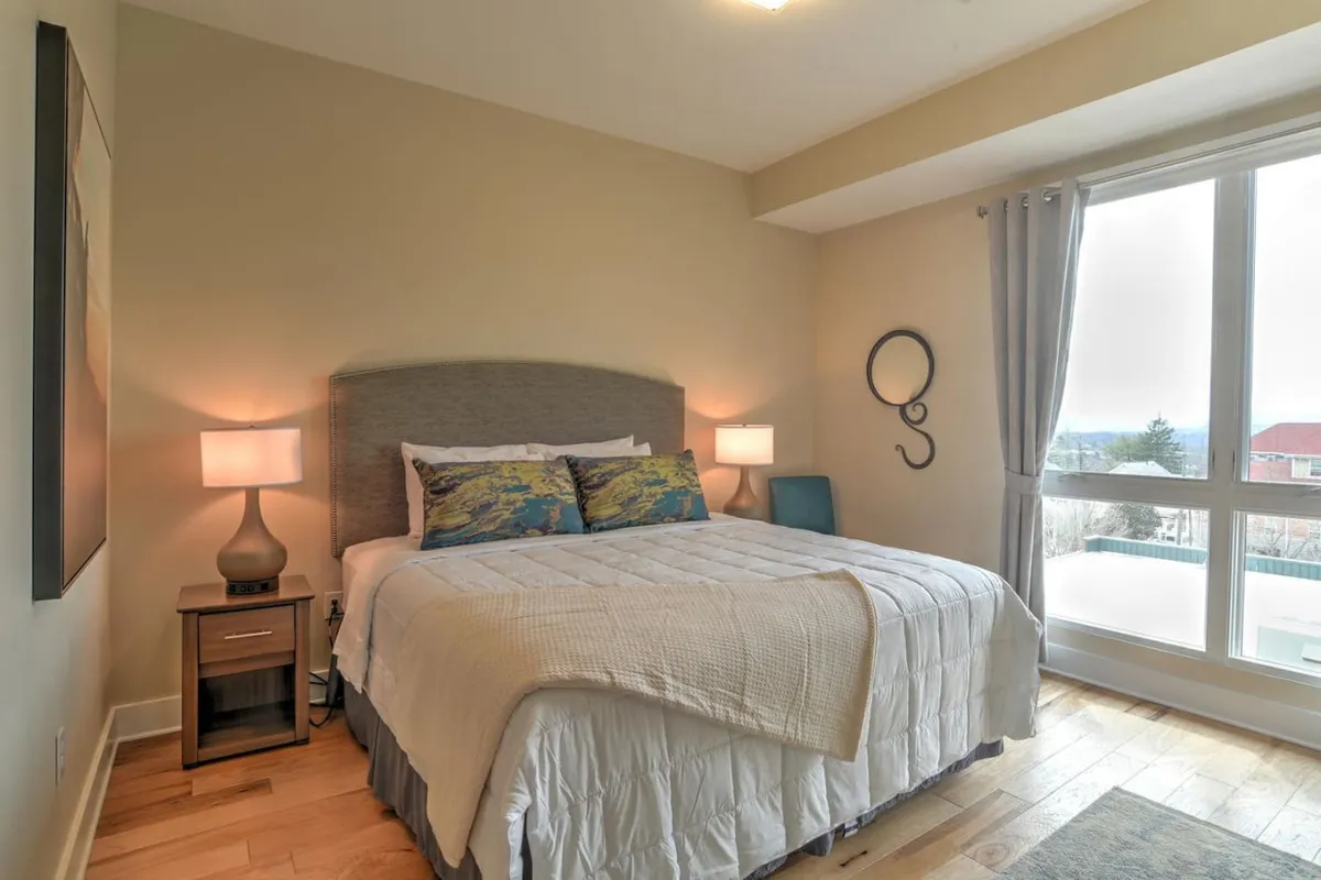 This image portrays a serene bedroom with a comfortable bed, a nightstand with a lamp, and a large window with curtains. Natural light filters into the room, evoking a peaceful and inviting atmosphere for guests and their pets to unwind after a day of exploring Asheville.