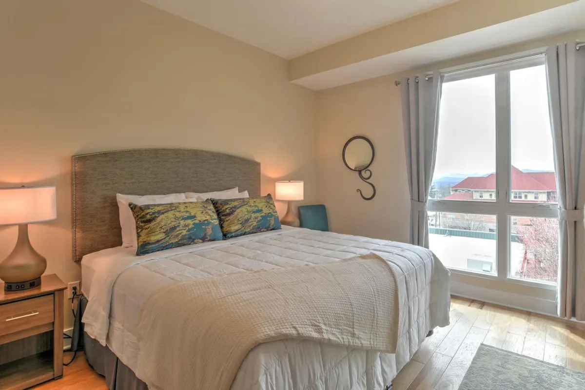 A tranquil bedroom scene featuring a cozy bed adorned with decorative pillows, a large window offering views beyond, and a nightstand with a lamp. The natural light and calm colors create an inviting space ideal for winding down with your pet.