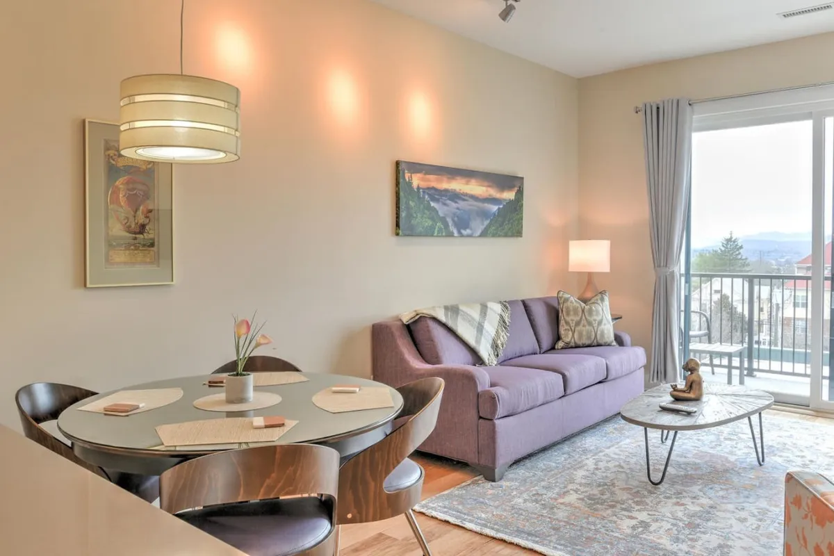 A sunlit open-plan living and dining area featuring a cozy sofa, a dining table set for two, eye-catching wall art, and a large window offering views of the sunset. The space exudes warmth and comfort, inviting relaxation and shared moments.