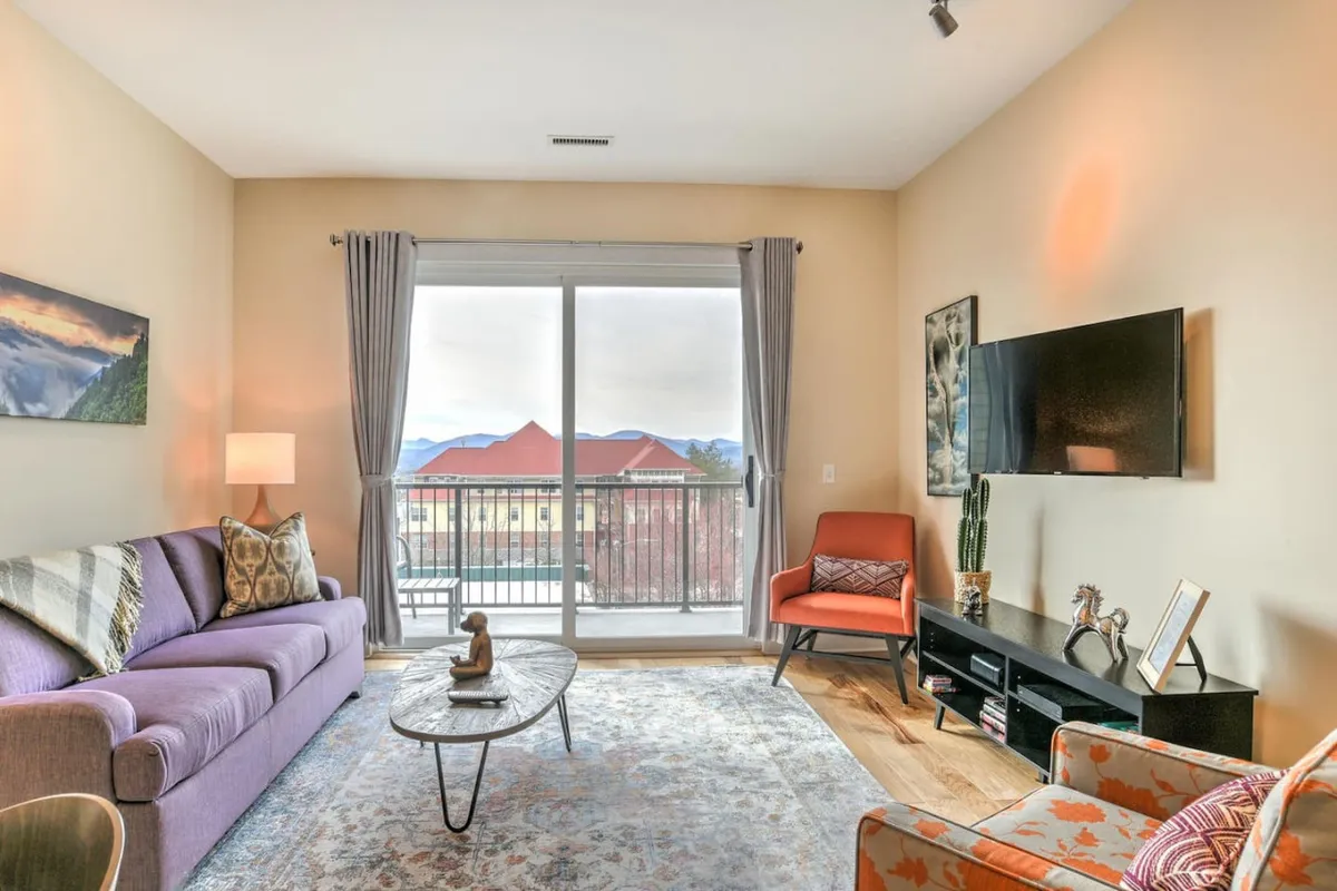 A comfortable living area highlighted by a plush purple pull-out sofa, a round coffee table, a large window offering mountain views, an orange accent chair, and a wall-mounted TV. The space is bathed in natural light and feels both welcoming and flexible for extra guests or relaxation.