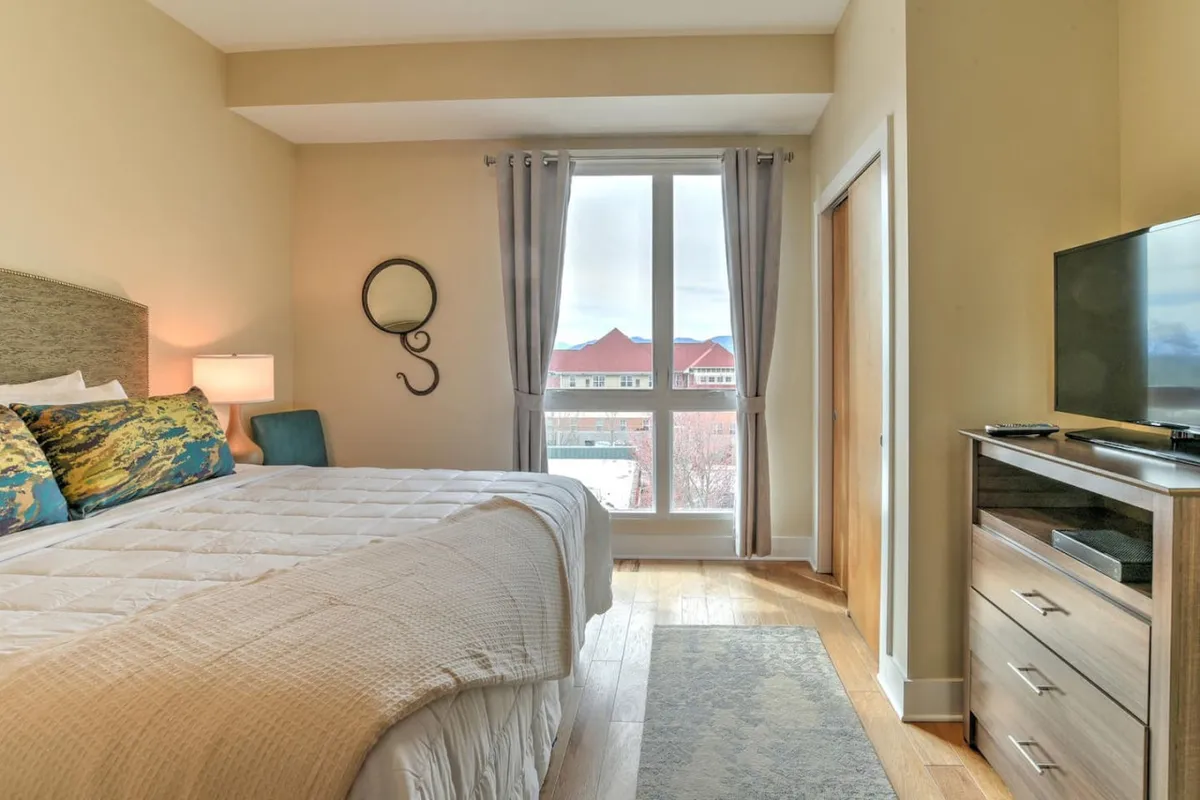 A spacious bedroom featuring a king-size bed with decorative pillows, a large window framing an inviting view, a TV on the dresser for entertainment, and a mirror that adds to the sense of space and light. The room exudes relaxation and comfort, with ample space and stylish touches.