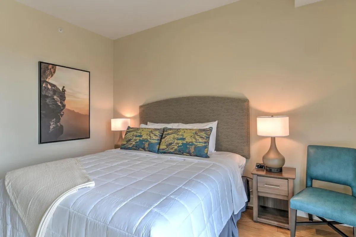 A serene bedroom with a plush king bed dressed in soft linens, decorative pillows adding a touch of style, a nightstand with a lamp for nighttime reading, and tasteful artwork above the headboard. The overall palette is calming, and the space feels private and inviting—perfect for unwinding after a busy day.